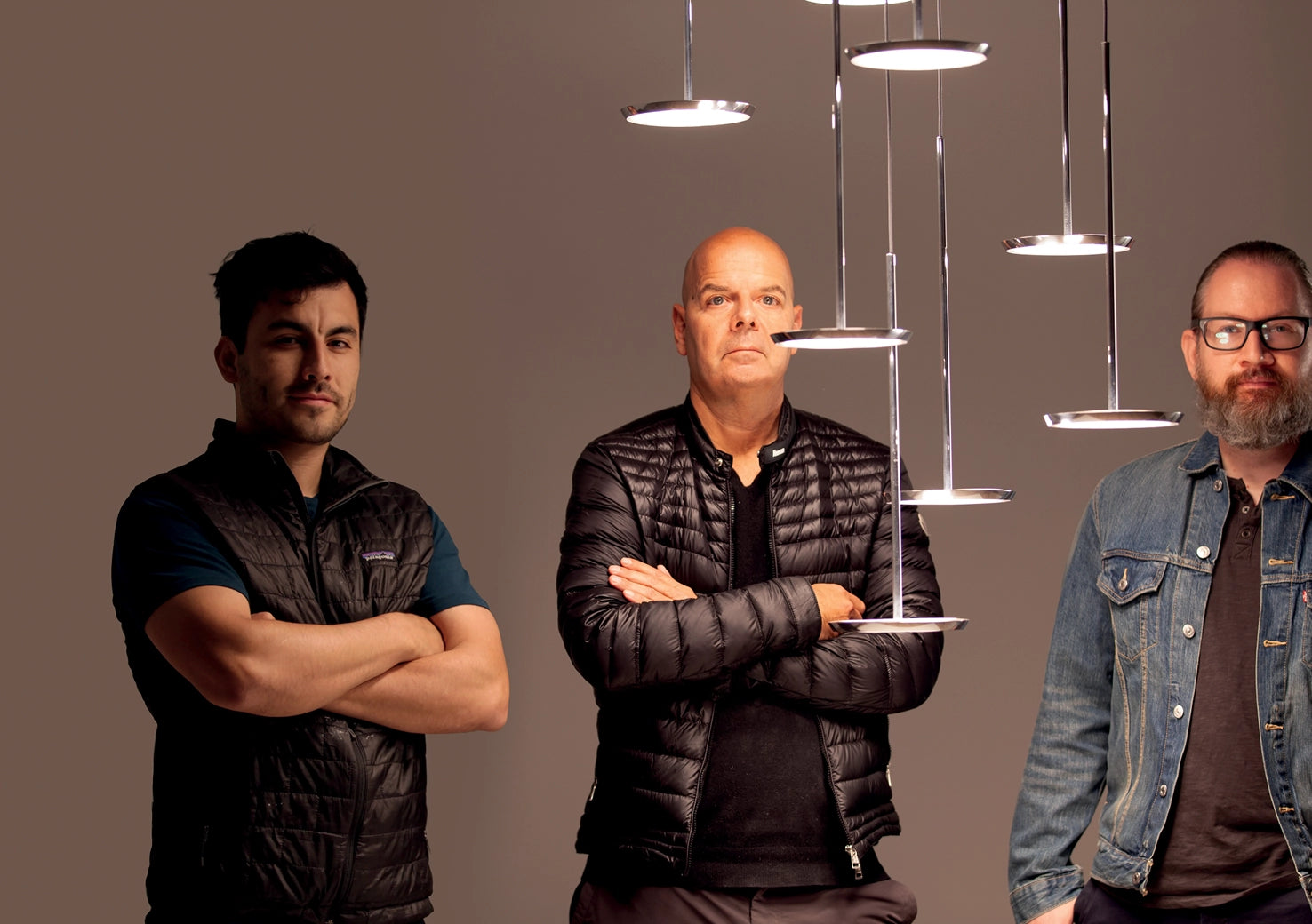 Three men standing in front of hanging lights against a brown background